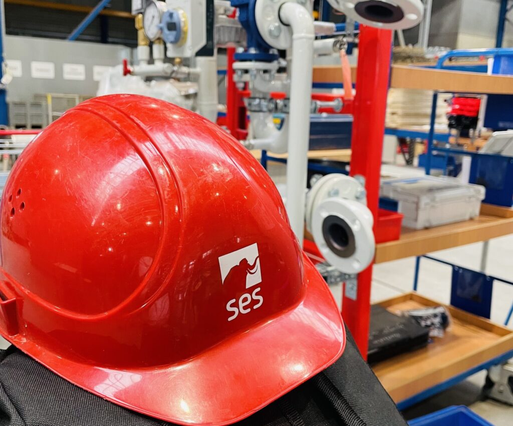 Das Bild zeigt einen Arbeitshelm von SES in rot in einer Werkstatt. Der Helm liegt auf einem Rucksack.
